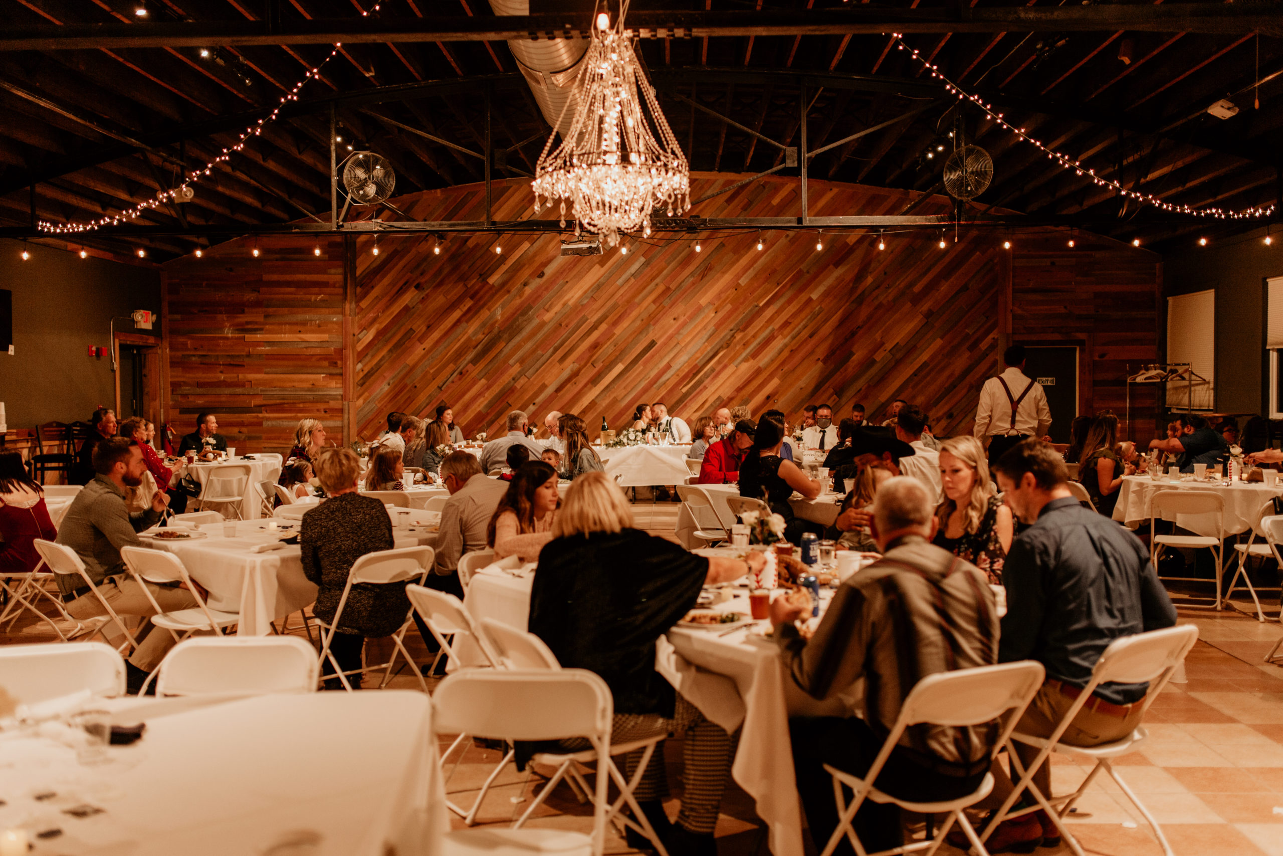 large wedding reception venue with wooden, plank walls and a large chandelier in the center of the room with string lights hung around